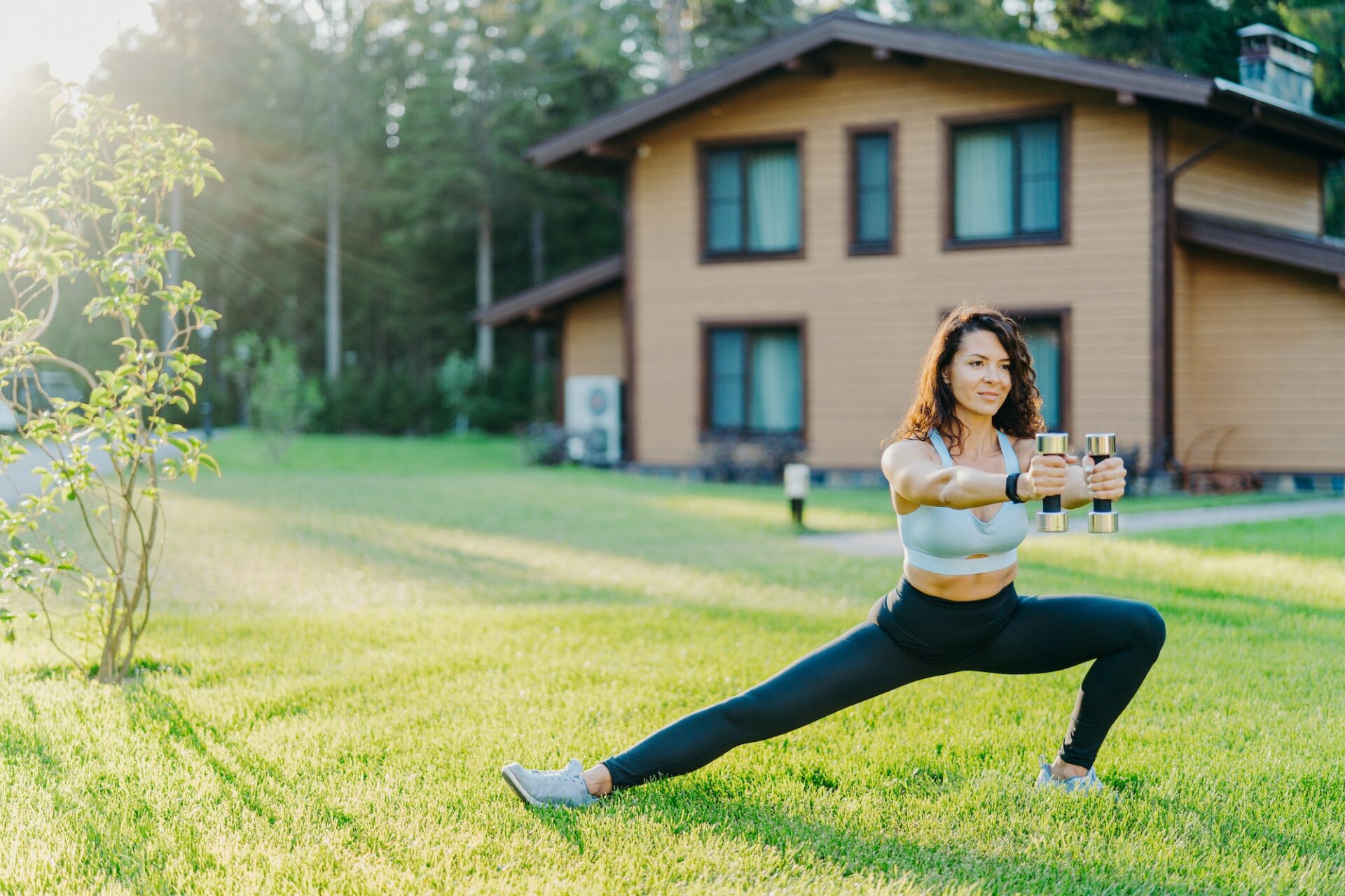 European sportswoman holds dumbbells and stretches legs, dressed in sport clothes